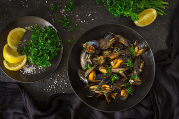Top view of cooked mussels in white wine, vegetables, herbs on black plate. Flat lay of seafood, lemon, salt crystals, parsley on dark background. High quality studio shot. 