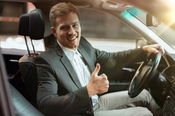 young handsome businessman driving luxury car looking satisfied, showing like sign, successful...