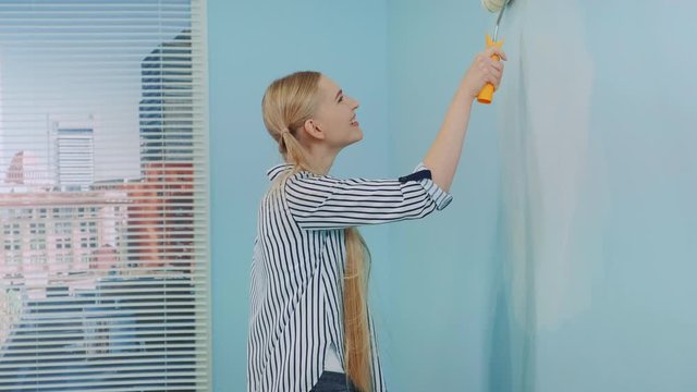 Woman makes repairs in the apartment dancing and smiling. She uses white paint and work with a roller. In the background there are skyscrapers at day.