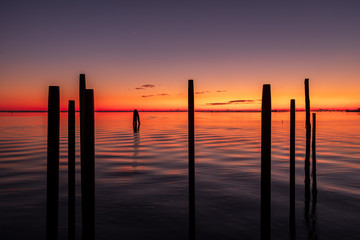 A silhouette of pylons in the sea at sunset with Venice in the background