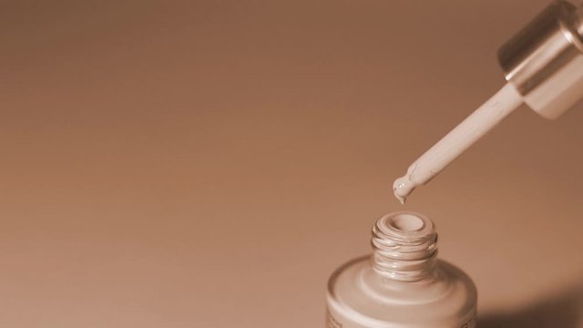 Foundation drips from a pipette into a glass bottle on a beige background. Monochrome video