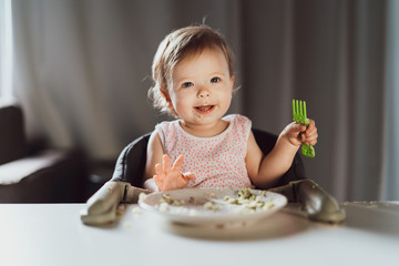 A nine-month-old smiling baby girl sits at a white table in a highchair and eats herself with a...