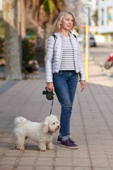 attractive middle-aged blond woman walking with fluffy white dog in summer city