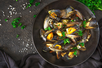 Top view of cooked mussels in white wine, vegetables, herbs on black plate. Flat lay of seafood, salt crystals, parsley on dark background. High quality studio photo.