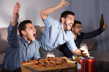 Happy men emotionally watching tv with beer and pizza at home