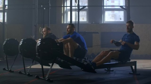 Three men work out together on rowing gym machines slow motion. Team training