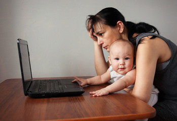 Young tired mother with one year old baby sits with a laptop. Work at home with children