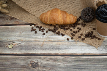 take away coffee cup , roast coffee bean and croissant on sack cloth in wooden background, homemade cafe concept 