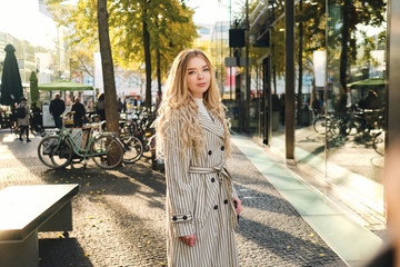 Beautiful blond girl in stylish trench coat walking through city street on sunny day