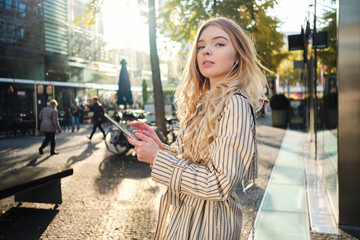 Fototapeta premium Beautiful blond girl in stylish trench coat with tablet walking through city street on sunny day