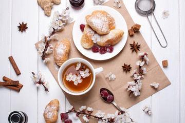 Spring card with a white cup of tea, spring branches, tasty croissants on a white wooden background in flat lay style