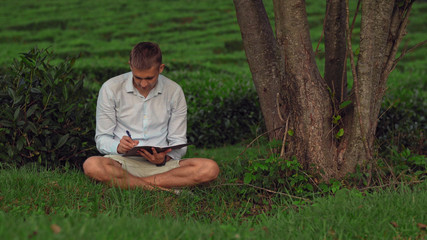 Naklejka na ściany i meble man sits under a tree on the grass and writes in a notebook.