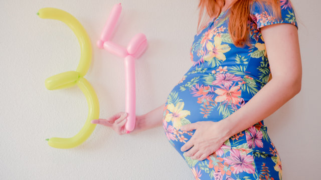 A close up photo of a pregnant young woman holding in her hands the week number thirty four (34) next to her belly in a beautiful dress. Photos of a pregnant woman in the third trimester expecting.