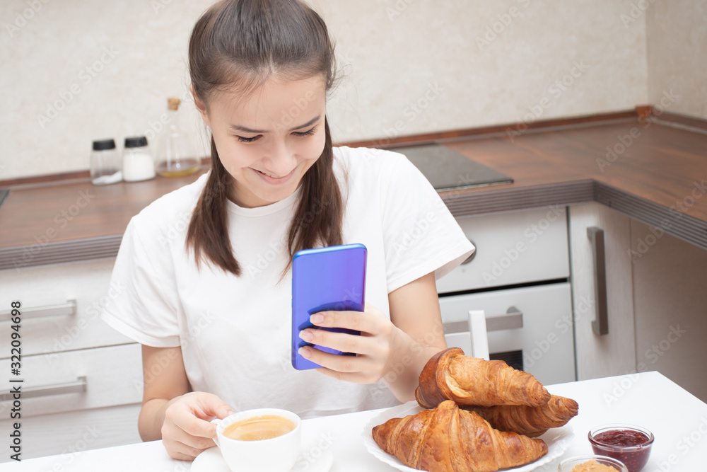 Wall mural a young girl looks at a smartphone, communicates on social networks at breakfast