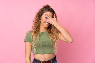 Young blonde woman with curly hair isolated on pink background covering eyes by hands and smiling