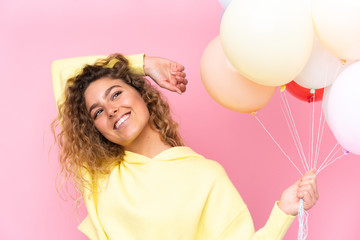 Young blonde woman with curly hair catching many balloons isolated on pink background