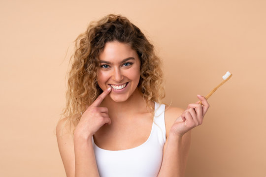 Young blonde woman with curly hair isolated on beige background with a toothbrush and happy expression