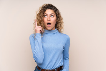 Young blonde woman with curly hair wearing a turtleneck sweater isolated on beige background thinking an idea pointing the finger up