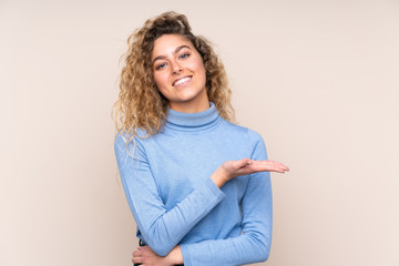 Young blonde woman with curly hair wearing a turtleneck sweater isolated on beige background presenting an idea while looking smiling towards