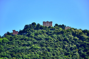 Fototapeta na wymiar Shore of Lake Como, region Lombardia, Italy - Three white-burgundy and beige buildings on top of a green mountain with trees, blue sky in the summer afternoon.