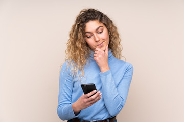 Young blonde woman with curly hair wearing a turtleneck sweater isolated on beige background thinking and sending a message