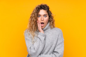 Young blonde woman with curly hair wearing a turtleneck sweater isolated on yellow background surprised and shocked while looking right
