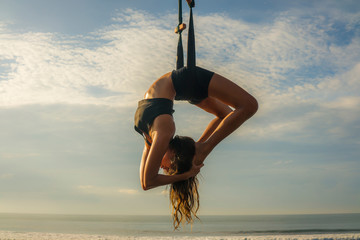 aero yoga beach workout - young attractive and athletic woman practicing aerial yoga exercise training acrobatic  body postures on blue sky over sea in Bali island