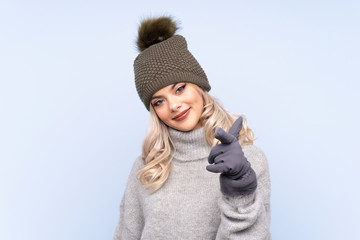 Young teenager girl with winter hat over isolated blue background points finger at you