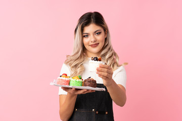 Teenager girl over isolated pink background holding mini cakes and offering it