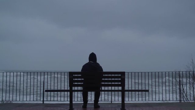 The man looks at the raging sea. A man is watching the storm. Silhouette of a man on the bench. Severe stormy sea. Big waves. Windy rainy weather. View of the dramatic ocean. The wind is blowing.