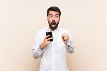 Young man with beard holding a mobile surprised and pointing front