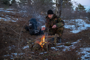 caucasian man in the camouflage sitting near the campfire in the winter forest, at night time