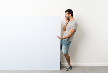 Young handsome man with beard holding a big blue empty placard with surprise and shocked facial expression