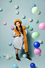 Fototapeta premium Little brunette asian girl in oversized shirt dress, brown beret and scarf, She posing with balloons on blue background.