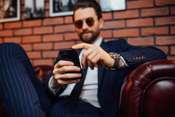 Handsome bearded man wearing  suit sitting on the leather sofa holding smartphone. Comfort and...