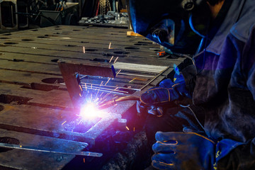 A welder in a workshop joins parts with a weld, sparks and smoke from welding.
