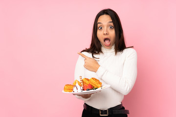 Young brunette woman holding waffles over isolated pink background surprised and pointing side