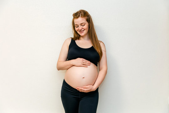 Image of pregnant woman in the black sport leggins and bra at thirty week (seven monthes) in the top and leggings, arms hugging her stomach. Pleasant expectation baby