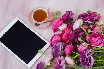 on a pink background a bouquet of ranunculus, a cup of tea and a tablet
