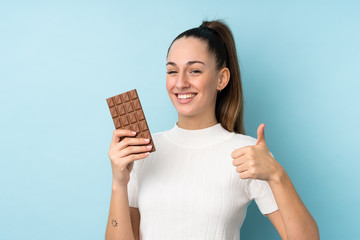 Young brunette woman over isolated blue background taking a chocolate tablet and with thumb up