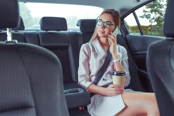 Foto op Plexiglas Beautiful girl secretary in business class of VIP taxi calling on phone, holding smartphone, fashionable and modern woman, cup of coffee with tea. Work on road, business meeting car. Meets passenger. © byswat