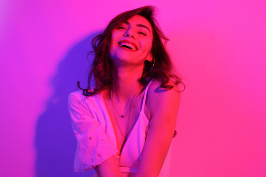 Brunette with long curled hair dancing in neon. A girl with professional makeup in red and blue light posing. The model laughs, smiles