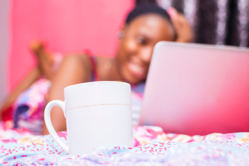 pretty african lady at home alone in her bedroom, watching a movie on her laptop pc, blurred, with coffee mug in focus