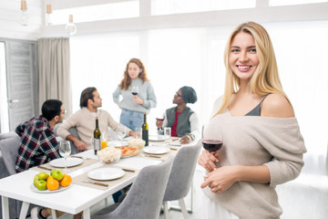 Portrait of young Caucasian woman with blond hair holding glass of wine looking at camera smiling, her friends chatting on background