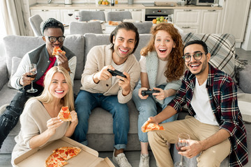 Group of five multi-ethnic friends having fun eating pizza and playing video game looking at camera, horizontal shot