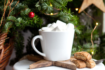 Coffee in a white cup with marshmallows. Morning festive coffee with traditional Italian cantuccini almond cookies. A cup of coffee on a background of green fir branches on a white stand.
