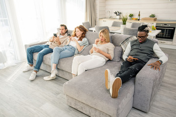 Obraz premium Horizontal shot of modern young people relaxing on sofa in cozy apartment using their smartphones
