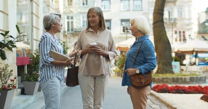 Three friendly good looking Caucasian old ladies standing in the city and talking. Cheerful beautiful grandmothers relaxing in the center of town in summer day. Senior women gossiping and having fun.