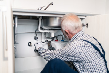 Naklejka na ściany i meble Senior man plumber working with plumbing tools on the kitchen. Renovation.