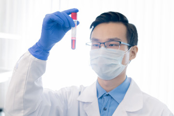 Unrecognizable Asian pharmacologist wearing eyeglasses, protective mask and gloves holding test tube with red liquid